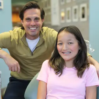 A smiling girl with braces in a pink shirt sits in a dental chair, while a man in a yellow shirt leans beside her in a brightly lit office.