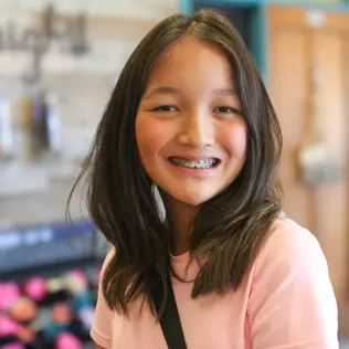 A smiling girl with long brown hair and braces, wearing a pink shirt. She stands indoors with colorful backgrounds, conveying a cheerful tone.