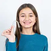 A smiling girl in a blue sweater holds a clear dental retainer, showcasing it proudly. The image conveys a sense of confidence and dental care.