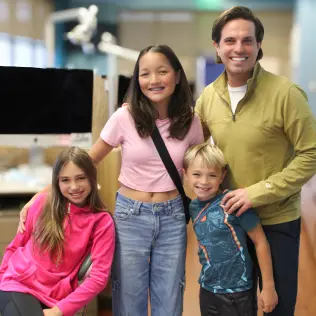 A smiling group of four, including a man and three children, pose together in a bright room. They convey a joyful, relaxed atmosphere.