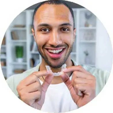 A smiling man holds a clear dental aligner, standing in a bright room with shelves in the background. He appears enthusiastic and content.