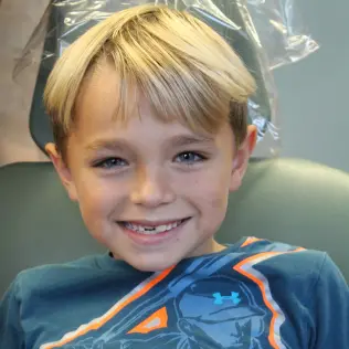 A smiling young boy with blonde hair sits in a chair wearing a blue shirt with a colorful design. The setting appears light and cheerful.