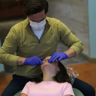 A dentist wearing gloves and a mask examines a patient's mouth in a relaxed dental office setting. The patient is reclining in the chair, evoking a calm atmosphere.